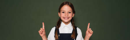 cheerful schoolgirl looking at camera while pointing with fingers near chalkboard, banner