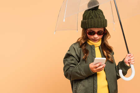 girl in trendy autumn outfit and sunglasses using smartphone under umbrella isolated on beigeの写真素材