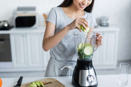 cropped view of smiling woman adding sliced zucchini into electric shaker