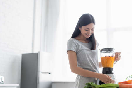 cheerful asian woman preparing vegetable smoothie for breakfast in kitchenの写真素材