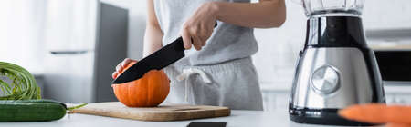 partial view of woman cutting fresh pumpkin near cucumbers and electric blender, bannerの写真素材