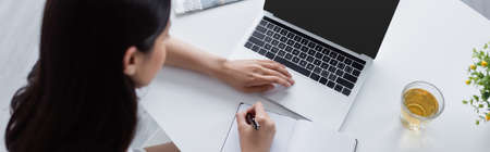 overhead view of woman writing in notebook near laptop and cup of tea on desk, banner