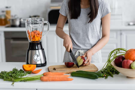partial view of woman cutting apple near raw vegetables and electric shakerの写真素材