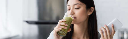pleased asian woman with smartphone drinking tasty smoothie with closed eyes, bannerの写真素材