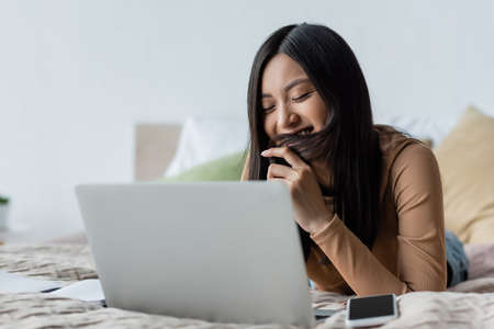 smiling asian woman covering mouth with hair near notebook in bedroomの写真素材