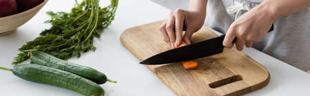 cropped view of woman cutting raw carrot on chopping board near fresh cucumbers on table, bannerの写真素材