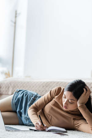 asian freelancer lying on floor in bedroom and writing in notebook near laptopの写真素材