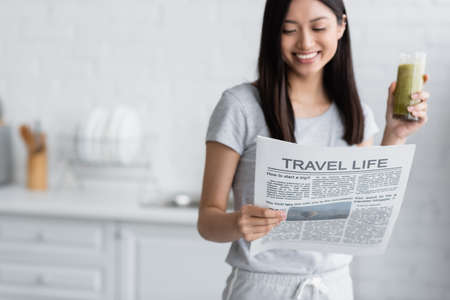 blurred asian woman with glass of fresh smoothie reading travel life newspaper in kitchenの写真素材
