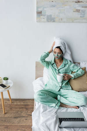 asian woman in clay mask and pajamas touching towel on head while sitting near laptop on bedの写真素材