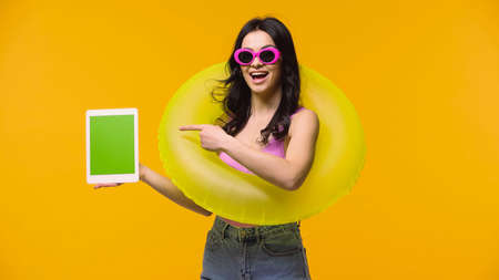 Excited woman in sunglasses and swim ring pointing at digital tablet isolated on yellow