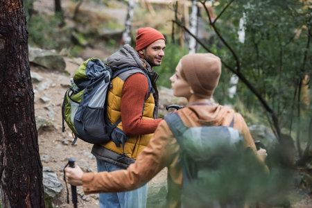 smiling man with backpack looking at woman trekking in forest on blurred foreground