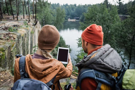back view of woman holding digital tablet with blank screen near boyfriend in forest near lakeの写真素材