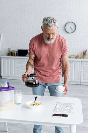 Mature man holding coffee pot and cup near breakfast, newspaper and smartphone in kitchenの写真素材