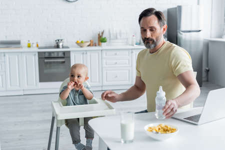 Mature man holding baby bottle near laptop, breakfast and daughter on high chairの写真素材