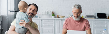 Smiling homosexual man holding daughter near partner using laptop in kitchen, banner