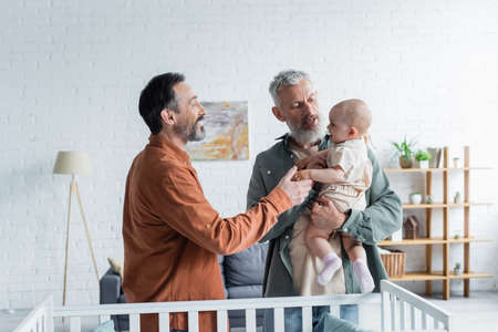Same sex family holding toddler daughter near baby crib