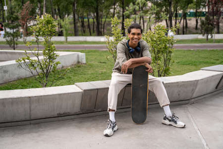 cheerful african american man smiling at camera while sitting on border bench with skateboardの写真素材
