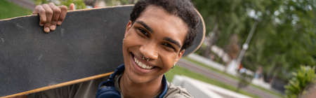 happy african american man with skateboard looking at camera outdoors, banner