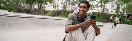 happy african american man with headphones and mobile phone sitting in skate park, bannerの写真素材