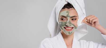 happy young woman in bathrobe removing clay mask with cotton pad isolated on gray, bannerの写真素材