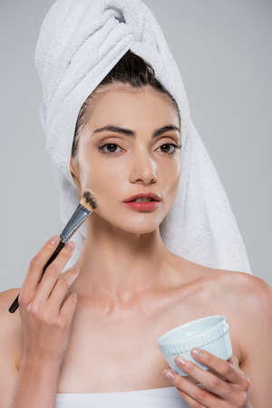 young woman with towel on head applying face mask with cosmetic brush isolated on grayの写真素材
