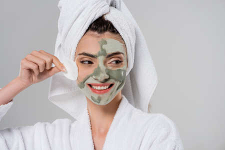 happy young woman in bathrobe removing clay mask with cotton pad isolated on grayの写真素材