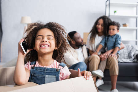 cheerful girl talking on mobile phone near blurred african american familyの写真素材