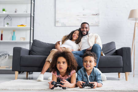 KYIV, UKRAINE - JANUARY 25, 2021: blurred african american couple smiling on sofa near kids gaming...