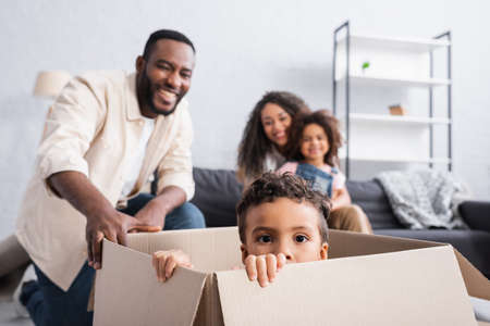 african american boy sitting in carton box near cheerful family on blurred background