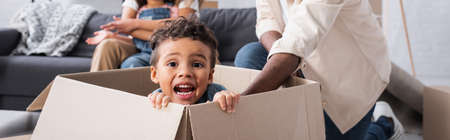 thrilled african american boy sitting in carton box near blurred family, banner