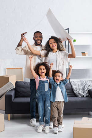 excited african american kids showing success gesture near parents with cardboard roof in new...