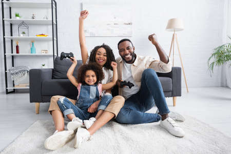 KYIV, UKRAINE - JANUARY 25, 2021: excited african american family showing triumph gesture while gaming on floor at homeの写真素材