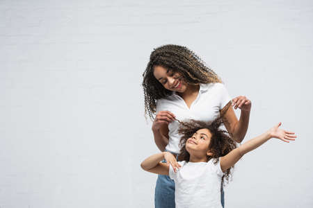joyful woman touching hair of curly african american daughter on gray