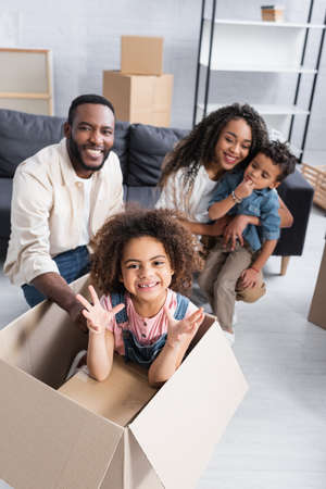 african american girl in carton box showing scaring gesture near blurred family in new apartment