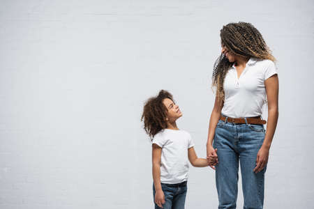 happy african american woman with daughter looking at each other while holding hands on gray