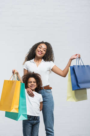 joyful african american woman with child holding colorful shopping bags on grayの写真素材
