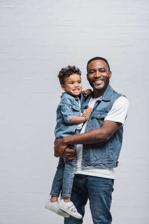 joyful african american man smiling at camera while holding son on grayの写真素材