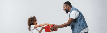 side view of african american man presenting gift to amazed daughter on gray, bannerの写真素材