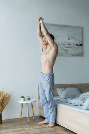 smiling man in striped pajama pants stretching with closed eyes in bedroom