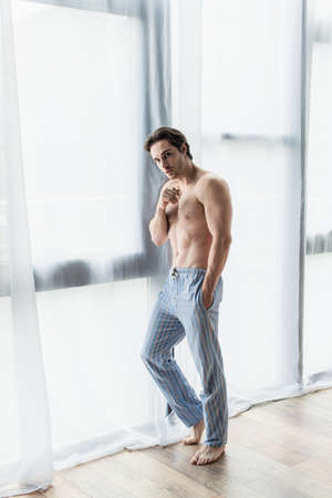 full length view of shirtless man in blue pajama pants near window at homeの写真素材