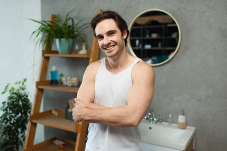 young man smiling at camera while standing with crossed arms in bathroomの写真素材