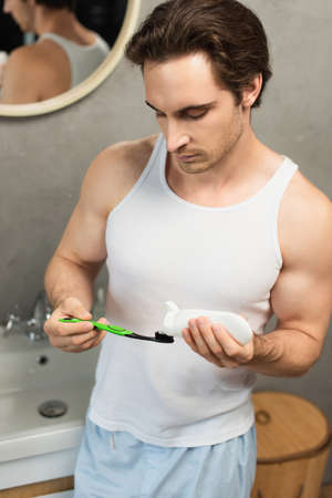 brunette man in white tank top applying toothpaste on toothbrush in bathroom