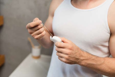 partial view of man in white tank top holding dental floss in bathroom