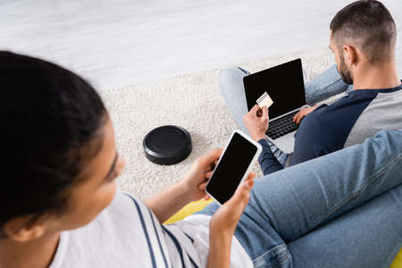 Robotic vacuum cleaner on carpet near interracial couple using devices and credit cardの写真素材