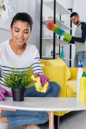 African american woman holding rag near plant and blurred boyfriend at homeの写真素材