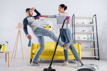Side view of african american woman with vacuum cleaner pointing at boyfriend with detergentの写真素材