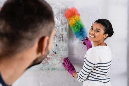 Smiling african american woman cleaning drawing with dust brush near blurred boyfriend