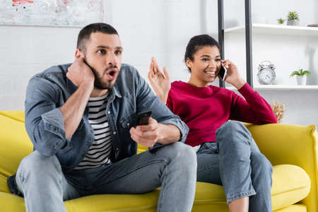 Smiling african american woman talking on mobile phone near excited boyfriend with remote controller