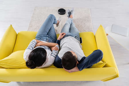 Overhead view of multiethnic couple sitting near devices and vacuum cleaner on couch