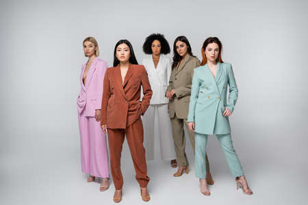 full length of interracial models in stylish suits posing with hands in pockets on gray
